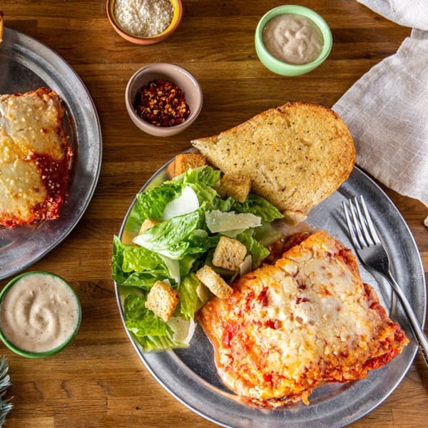Two plates of DaVinci's Pizzeria lasagna with Caesar salad, garlic bread, and condiments on a wooden table.