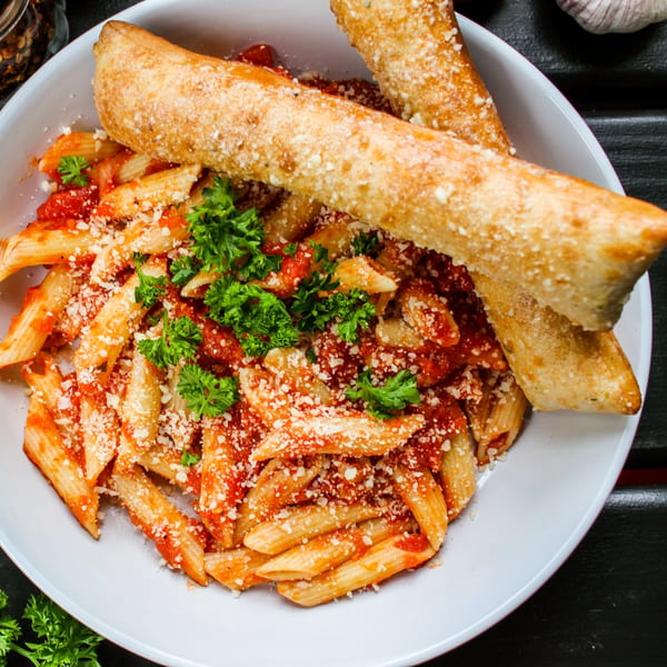 Penne pasta with red sauce, parmesan cheese, parsley, and two breadsticks in a white bowl.