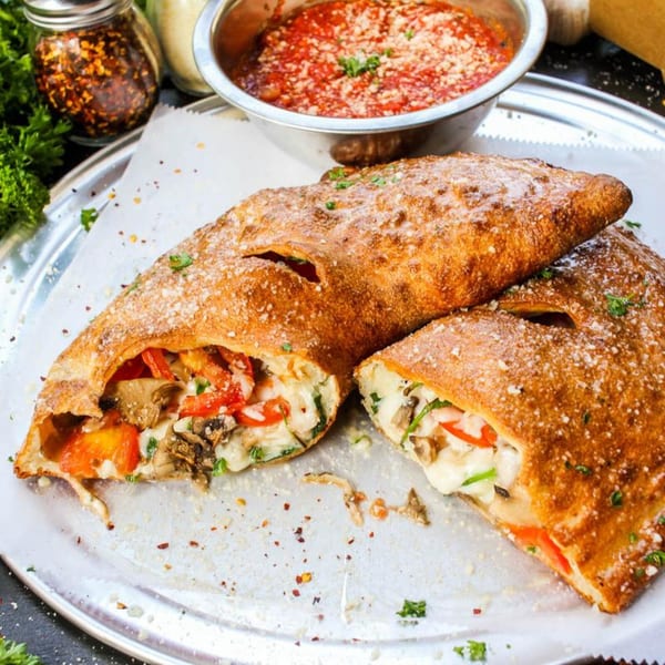 Two golden-brown calzones, one sliced open revealing melted cheese, mushrooms, and red peppers, with marinara sauce.