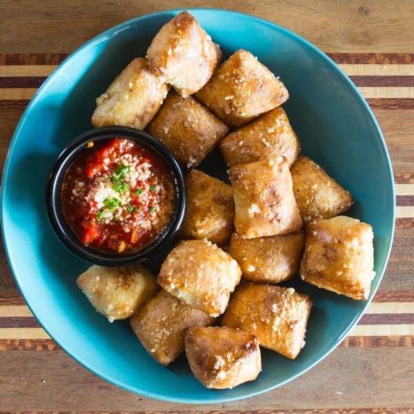 Golden-brown garlic knots dusted with Parmesan cheese and parsley, served with marinara sauce in a blue bowl.