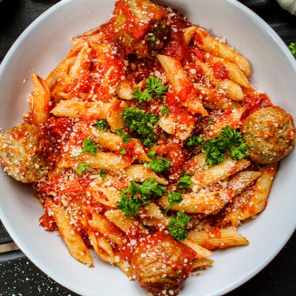 Penne pasta with meatballs, marinara sauce, parmesan cheese, and fresh parsley in a white bowl.